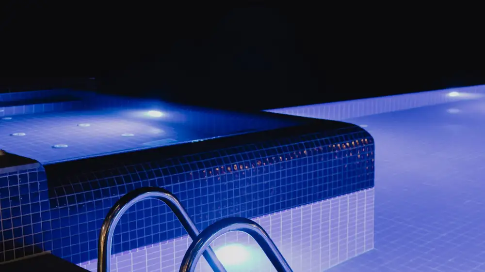 A pool lit from within at night beside a steel handrail